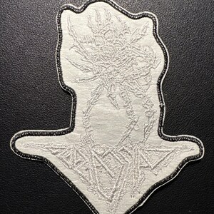 以下が含まれることがあります： 黒い縁取りのある白い刺繍パッチ。パッチは、中央の図と幾何学的な形状を備えた詳細な抽象的なデザインを特徴としています。パッチは、おそらく衣類またはアクセサリー用です。