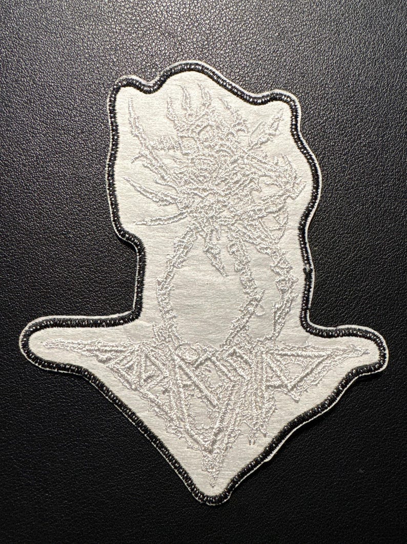 以下が含まれることがあります： 黒い縁取りのある白い刺繍パッチ。パッチは、中央の図と幾何学的な形状を備えた詳細な抽象的なデザインを特徴としています。パッチは、おそらく衣類またはアクセサリー用です。