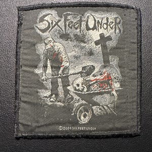 Puede incluir: Un parche en blanco y negro con el texto "Six Feet Under" en una fuente gótica. La imagen muestra una figura empujando una carretilla con una calavera y restos. Una cruz está en el fondo. El texto "©2008 SIX FEET UNDER" está en la parte inferior.
