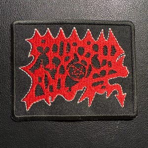 May include: A rectangular black patch with a red, stylized design and white stitching. The red design features jagged edges and an intricate pattern. The patch is likely for clothing or accessories.