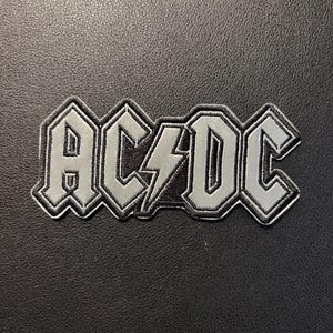 Könnte beinhalten: Ein silber-schwarzer Stickaufnäher mit dem Schriftzug AC/DC und einem Blitzsymbol zwischen den Buchstaben. Das Design hat eine metallisch silberne Oberfläche für die Buchstaben und den Blitz, umrandet von einem dicken schwarzen Rand.
