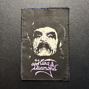May include: A rectangular black fabric patch featuring a white and light purple graphic of a face with heavy theatrical makeup. Below the face, the text King Diamond is printed in a stylized light purple font with a small cross symbol above the i.