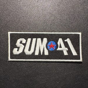 May include: Black rectangular patch with the text "SUM 41" in white, embroidered lettering. The "O" in "SUM" features a blue circle with a red star. The patch has a white border.