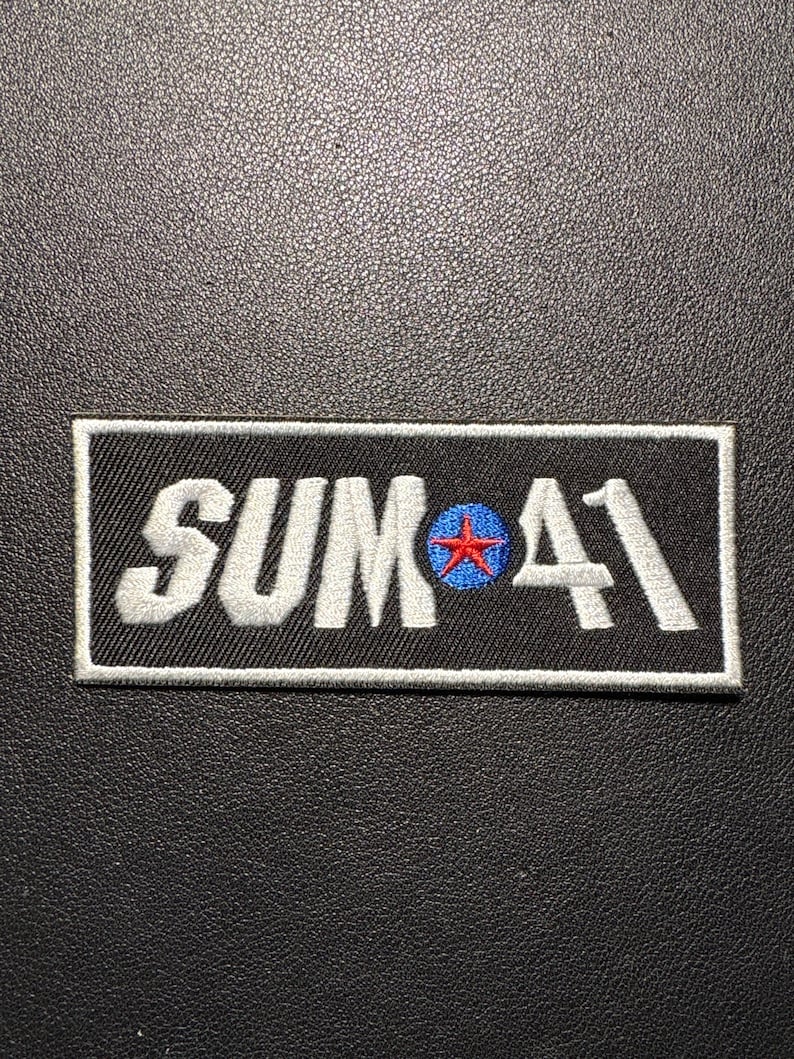 May include: Black rectangular patch with the text "SUM 41" in white, embroidered lettering. The "O" in "SUM" features a blue circle with a red star. The patch has a white border.
