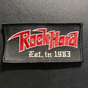 Könnte beinhalten: Ein rechteckiger, schwarz bestickter Aufnäher mit dem roten Schriftzug "RockHard" in einer stilisierten Schriftart und dem darunter platzierten weißen Text "Est. in 1983". Der Aufnäher hat einen schwarzen Stickrand und einen strukturierten Stoffhintergrund.