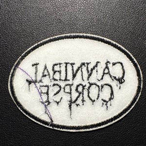 May include: Oval patch featuring the text "CANNIBAL CORPSE" in black, stylized lettering. The patch has a white background with a black border. The patch is on a black surface.
