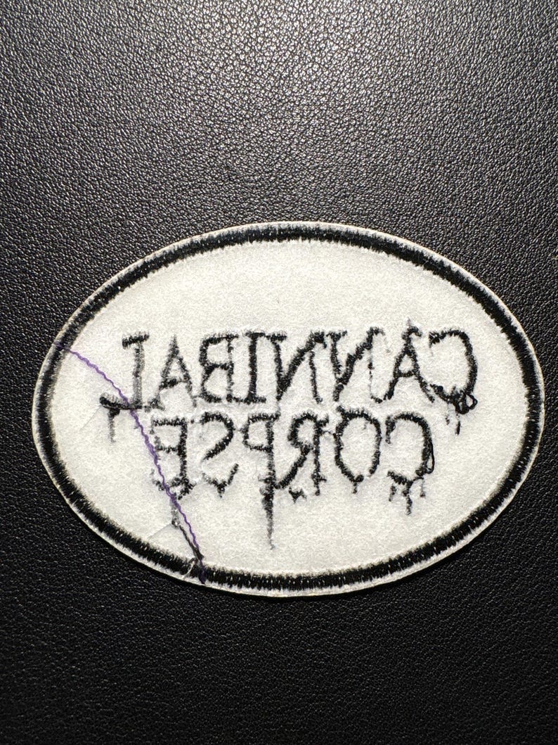 May include: Oval patch featuring the text "CANNIBAL CORPSE" in black, stylized lettering. The patch has a white background with a black border. The patch is on a black surface.