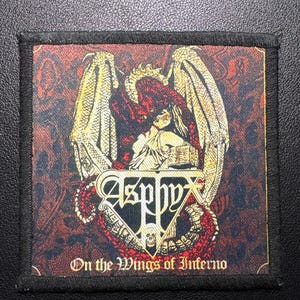 Puede incluir: Parche cuadrado con borde negro. El diseño presenta una criatura alada, el nombre de la banda "Asphyx" y el texto "On the Wings of Inferno". La paleta de colores incluye rojo, dorado y negro.