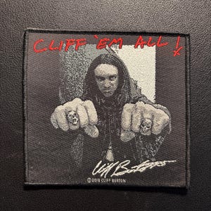 Può includere: Toppa in bianco e nero con il ritratto di una persona con i capelli lunghi, i pugni chiusi e anelli a teschio. Il testo "CLIFF 'EM ALL!" è ricamato in rosso. È visibile anche la firma "Cliff Burton".