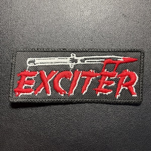 Puede incluir: Un parche rectangular bordado con fondo negro. La palabra EXCITER está bordada en rojo con un contorno blanco. Sobre el texto, se representa una navaja automática blanca con una punta roja en la hoja.