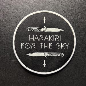 Könnte beinhalten: Ein runder, schwarzer Aufnäher mit weißem Rand. Der Aufnäher zeigt zwei Messer, ein Kreuz und den Text "HARAKIRI FOR THE SKY" in Weiß. Die Messer sind oberhalb und unterhalb des Textes positioniert, mit einem Kreuz oberhalb und unterhalb der Messer.