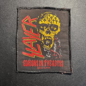 May include: A black square patch featuring the word "SLAYER" in red, with a yellow skull graphic. The text "SEASONS IN THE ABYSS" is below the skull. The patch is made of fabric.