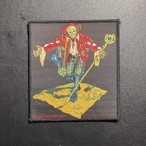 Puede incluir: Parche cuadrado bordado que representa una figura esquelética con una túnica roja y dorada, sosteniendo un bastón con una gema verde. La figura está sobre una plataforma amarilla con una rosa. Texto: "©1995 THE ALCHEMY CARDS".