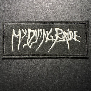Könnte beinhalten: Ein rechteckiger, dunkelgrauer Aufnäher mit dem weißen Text "MY DYING BRIDE" aufgestickt. Der Aufnäher hat einen schwarzen Rand und ist vor einem dunklen Hintergrund platziert. Der Text ist in einer stilisierten, gotischen Schriftart.