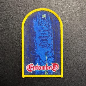 May include: A rectangular patch with a yellow border and a blue interior. The blue field features a tombstone design with the words "REST IN FESTERING SLIME" and the band name "Entombed" in red script.
