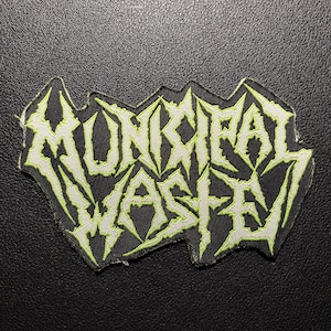 Può includere: Una toppa in bianco e nero con la scritta "MUNCIPAL WASTE" in un font verde neon frastagliato. La toppa ha un'estetica punk rock usurata.