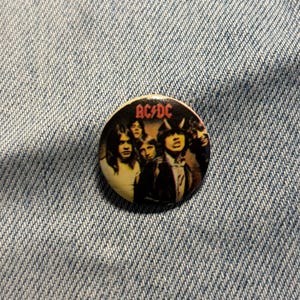 Könnte beinhalten: Ein runder Ansteckbutton mit einem Vintage-Foto der Rockband AC/DC. Die Bandmitglieder sind in einem Gruppenporträt zu sehen, wobei das rote AC/DC-Logo oben aufgedruckt ist. Der Button hat eine glänzende Oberfläche und einen metallischen Rand.