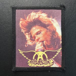 Könnte beinhalten: Aerosmith-Band-Patch mit einem Porträt eines Sängers mit langen, rotbraunen Haaren, der in ein Mikrofon singt. Der Patch hat das Logo der Band mit Flügeln und den Namen der Band in goldfarbenen Buchstaben. Der Patch ist rechteckig mit einem schwarzen Rand.