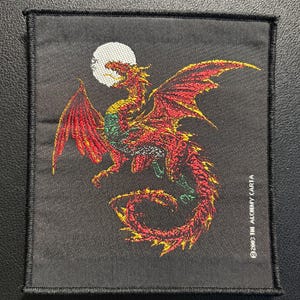 Könnte beinhalten: Ein rechteckiger, schwarzer, bestickter Aufnäher mit einem roten und orangefarbenen Drachen mit grünen Akzenten vor einem weißen, kreisförmigen Mond. Der Text 2003 THE ALCHEMY CARTA ist in Weiß am unteren rechten Rand aufgedruckt.