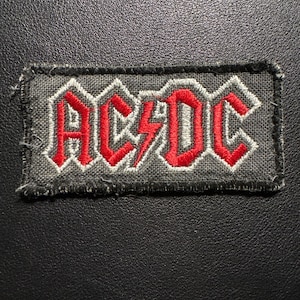 May include: Rectangular embroidered patch with the band name "AC/DC" in red, outlined in white, on a grey background. The patch has a black border. Suitable for application on clothing or accessories. The design includes a lightning bolt.
