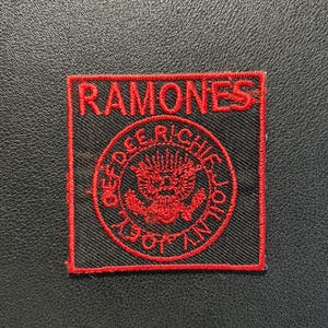 Könnte beinhalten: Ein quadratischer gestickter Aufnäher mit schwarzem Hintergrund und rotem Rand. Der Text RAMONES steht oben. Darunter befindet sich ein kreisförmiges Emblem mit einem Adler-Motiv, umgeben von den Namen DEE DEE, RICHIE, JOEY und JOHNNY in roter Stickerei.