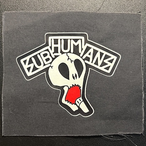 May include: A rectangular dark gray fabric patch featuring a white graphic of a skull with a red mouth area. Above the skull, the word SUBHUMANS is printed in a bold, stylized white font with black outlines, set against a black background.