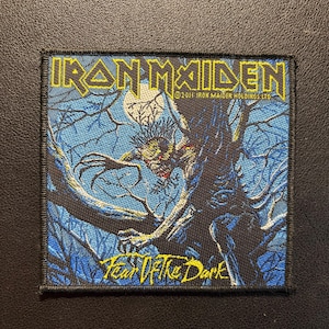 Peut inclure: Écusson brodé carré avec le logo Iron Maiden en jaune et le texte "Fear of the Dark". Le motif représente une créature dans un arbre sur fond bleu. L'écusson a une bordure noire.