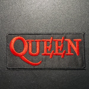 Könnte beinhalten: Ein rechteckiger, schwarzer Aufnäher mit dem Wort "QUEEN" in roter Stickerei. Der Aufnäher hat einen schwarzen Rand und befindet sich auf einem strukturierten, dunklen Hintergrund.