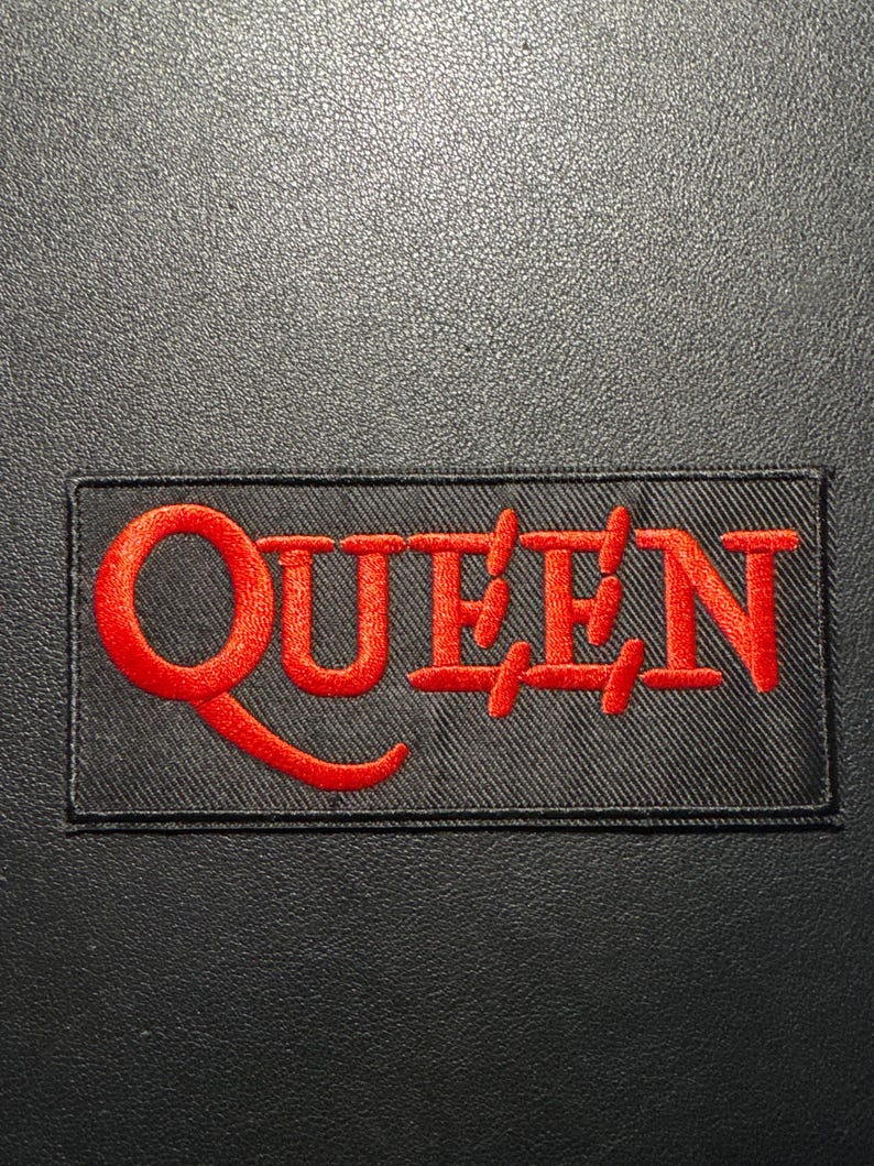 Puede incluir: Un parche negro rectangular con la palabra "QUEEN" bordada en rojo. El parche tiene un borde negro y est&aacute; sobre un fondo oscuro y texturizado.