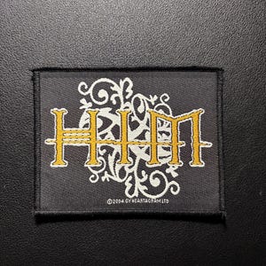 Könnte beinhalten: Schwarzer rechteckiger Aufnäher mit den Buchstaben "HIM" in Gold und einem weißen Zierdesign. Der Text "©2004 BY HEARTAGRAM LTD" befindet sich unten. Der Aufnäher liegt auf einer schwarzen Oberfläche.