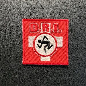 Op de afbeelding: Een vierkante geborduurde patch met een felrode achtergrond. De tekst D.R.I. is in het wit aan de bovenkant geborduurd. Onder de tekst staat een witte cirkel met een zwart stokfiguurtje in een hardloophouding, op een witte kruisvorm.