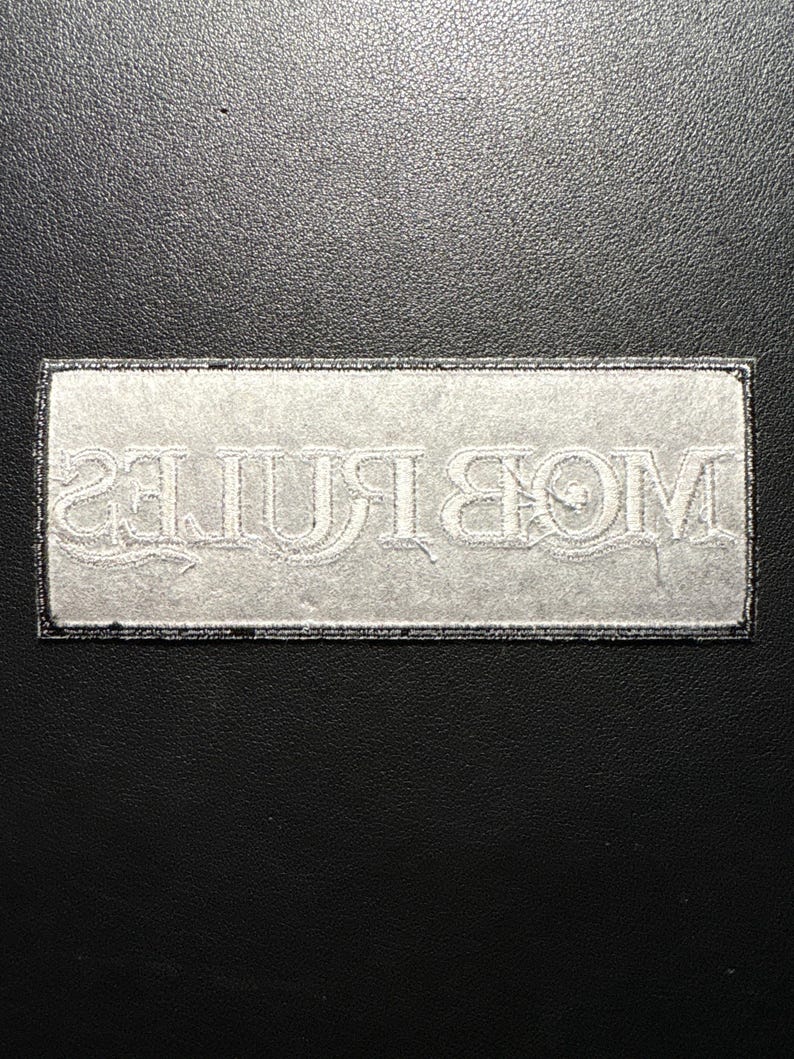 May include: A rectangular patch with the word "MOB RULES" embroidered in white thread. The patch has a black border and is set against a black background. The text is in a stylized font.