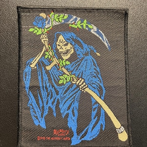Pode incluir: Um emblema retangular bordado em preto com uma figura de esqueleto vestindo um manto azul com capuz, segurando uma foice envolta em vinhas verdes com uma rosa azul. O texto "ALCHEMY GOTHIC" e "©1995 THE ALCHEMY CARTA" está em vermelho.