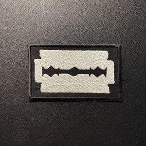 May include: A rectangular embroidered patch featuring a white razor blade design centered on a solid black background. The patch has a stitched border and displays the iconic shape of a double-edged razor blade with a central cutout detail.
