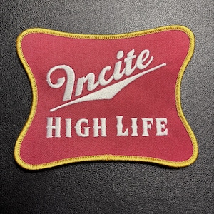 Puede incluir: Un parche rojo con borde amarillo con la palabra "Incite" en escritura blanca sobre "HIGH LIFE" en letras mayúsculas blancas. El parche tiene una forma única y es probablemente un artículo para planchar o coser.