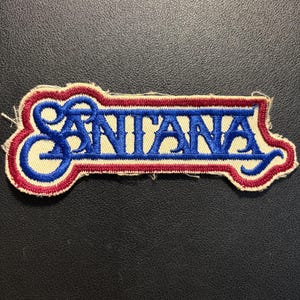 Può includere: Una toppa in tessuto ricamato che presenta la parola SANTANA in lettere blu in grassetto. Il testo è delineato da un bordo rosso spesso, ulteriormente circondato da uno sfondo color crema. La toppa ha una forma irregolare distintiva.