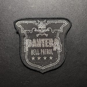 Könnte beinhalten: Schwarz-silberner, gestickter Aufnäher mit dem Text "PANTERA HELL PATROL" und einem Totenkopf- und gekreuzten Knochen-Design. Der schildförmige Aufnäher hat einen silbernen Rand und vier Sterne am unteren Rand. Zum Aufnähen auf Kleidung oder Taschen.