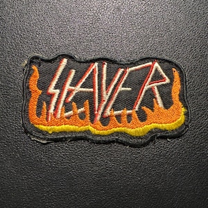 Könnte beinhalten: Gestickter Aufnäher mit dem Wort "SLAYER" in weißen und roten Blockbuchstaben auf schwarzem Hintergrund. Der Aufnäher ist mit orangefarbenen und gelben Flammen umrandet.