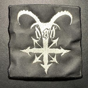 Könnte beinhalten: Schwarzer quadratischer Aufnäher mit weißem Stickdesign. Das Design zeigt einen Ziegenkopf mit umgekehrten Hörnern über einem Kreuz mit nach außen zeigenden Pfeilen. Der Aufnäher hat einen schwarzen Rand.