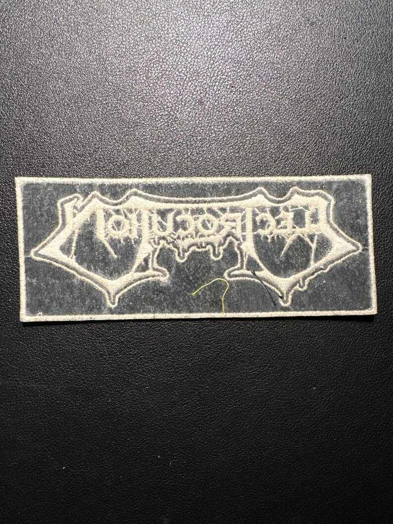 May include: A rectangular patch featuring the word "MORBID" in a gothic font. The text is white with a darker outline, set against a grey background. The patch has a white border.