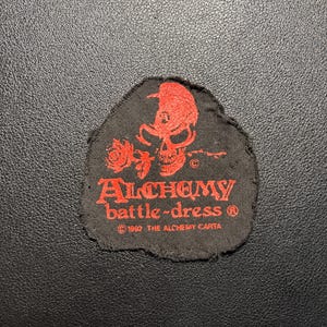 Könnte beinhalten: Ein schwarzer Stoffaufnäher mit einer roten Grafik eines Totenkopfes und einer Rose. Der Text lautet ALCHEMY battle-dress und 1992 THE ALCHEMY CARTA. Der Aufnäher hat ausgefranste Ränder und eine unregelmäßige Form mit einer flachen Unterkante.