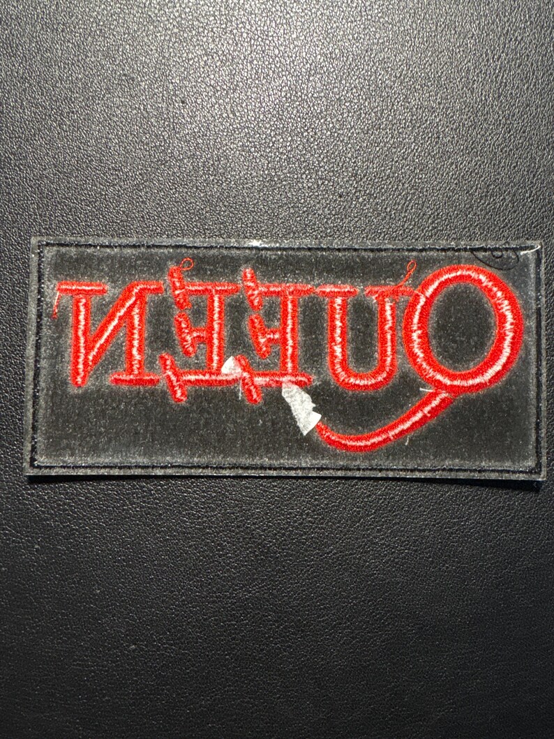 Puede incluir: Parche bordado con la palabra "QUEEN" en hilo rojo sobre un fondo rectangular negro. El parche tiene un borde blanco y est&aacute; sobre una superficie negra texturizada.