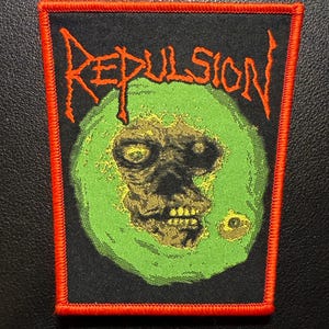 May include: A black patch with an orange border featuring the word "REPULSION" in orange, jagged lettering. The patch has a green circle with a zombie-like skull in the center. The skull is a mix of brown and yellow.