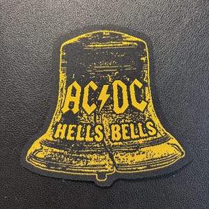 Könnte beinhalten: Ein schwarzer, glockenförmiger Aufnäher mit einer gelben, abgenutzten Grafik. Das Design zeigt den Schriftzug AC/DC mit einem Blitz in der Mitte und darunter die Worte HELLS BELLS in einer fetten, gelben Schrift in Großbuchstaben.