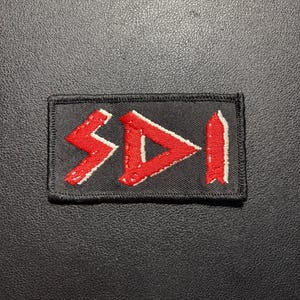 Puede incluir: Un parche rectangular bordado en negro que presenta las letras rojas S, D e I en una fuente estilizada. Las letras están delineadas con hilo blanco y todo el parche tiene un borde cosido en negro, creando un diseño audaz y de alto contraste.