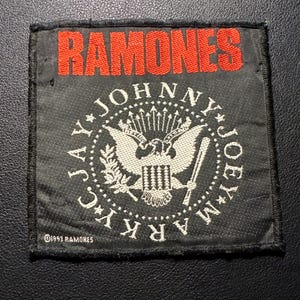 Könnte beinhalten: Ein schwarzer rechteckiger Aufnäher mit dem Wort "RAMONES" in Rot über einem kreisförmigen Emblem. Das Emblem zeigt einen Adler, Pfeile und die Namen "JOHNNY", "JOEY", "MARK" und "JAY". Der Text "©1993 RAMONES" befindet sich unten.