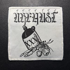 May include: A square white fabric patch with a black embroidered design featuring a hermit crab emerging from a jug labeled with three X's. Above the crab and jug, the text "WREHYUST" is embroidered in a stylized black gothic font.