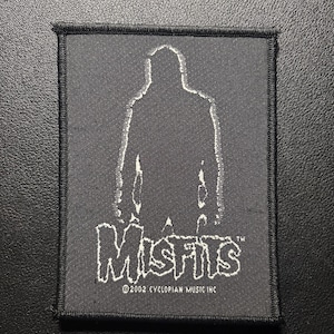 Può includere: Toppa quadrata nera con una silhouette bianca di una figura e la parola "Misfits" in un font stilizzato. La toppa include anche il testo "©2002 CYCLOPIAN MUSIC INC". La toppa ha un bordo nero.