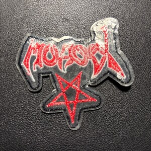 Op de afbeelding: Geborduurde patch met het woord "MOTOR" in rode, gestileerde letters met een zwarte omlijning. Onder het woord staat een rode pentagram. De patch heeft een zwarte achtergrond en een witte rand.
