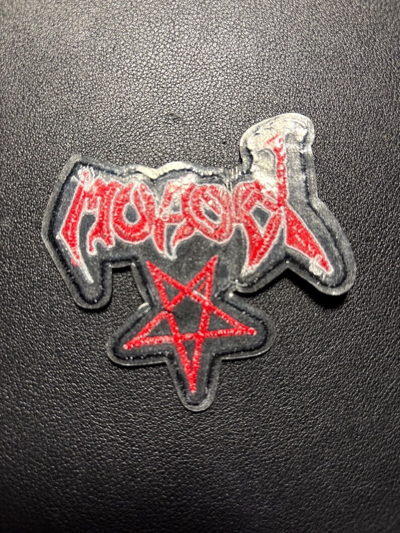 Op de afbeelding: Geborduurde patch met het woord "MOTOR" in rode, gestileerde letters met een zwarte omlijning. Onder het woord staat een rode pentagram. De patch heeft een zwarte achtergrond en een witte rand.